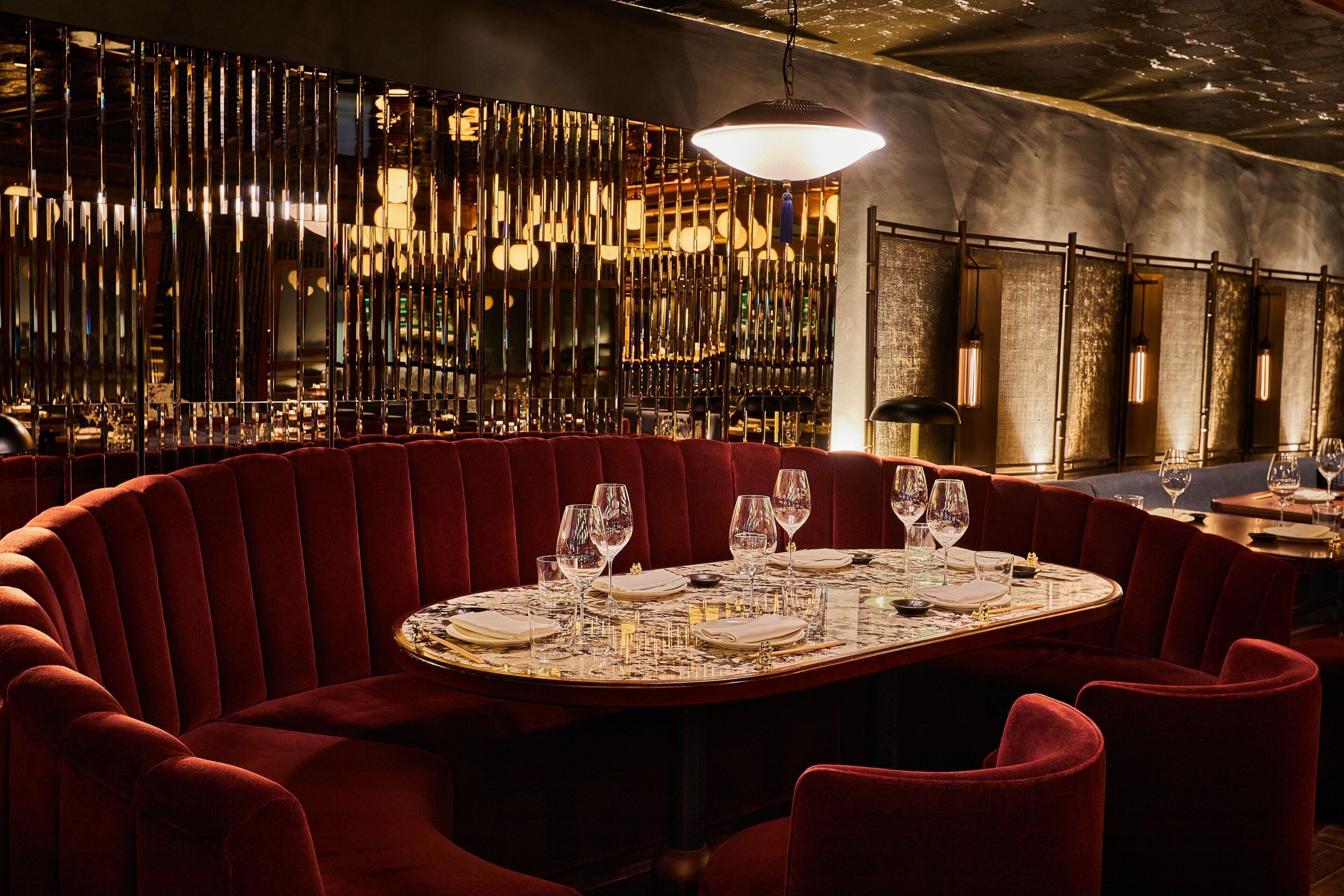 A round table with red upholstered chairs, set for dining, in a dimly lit room with a decorative wall and hanging light fixtures.