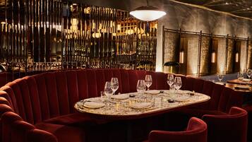 A round table with red upholstered chairs, set for dining, in a dimly lit room with a decorative wall and hanging light fixtures.
