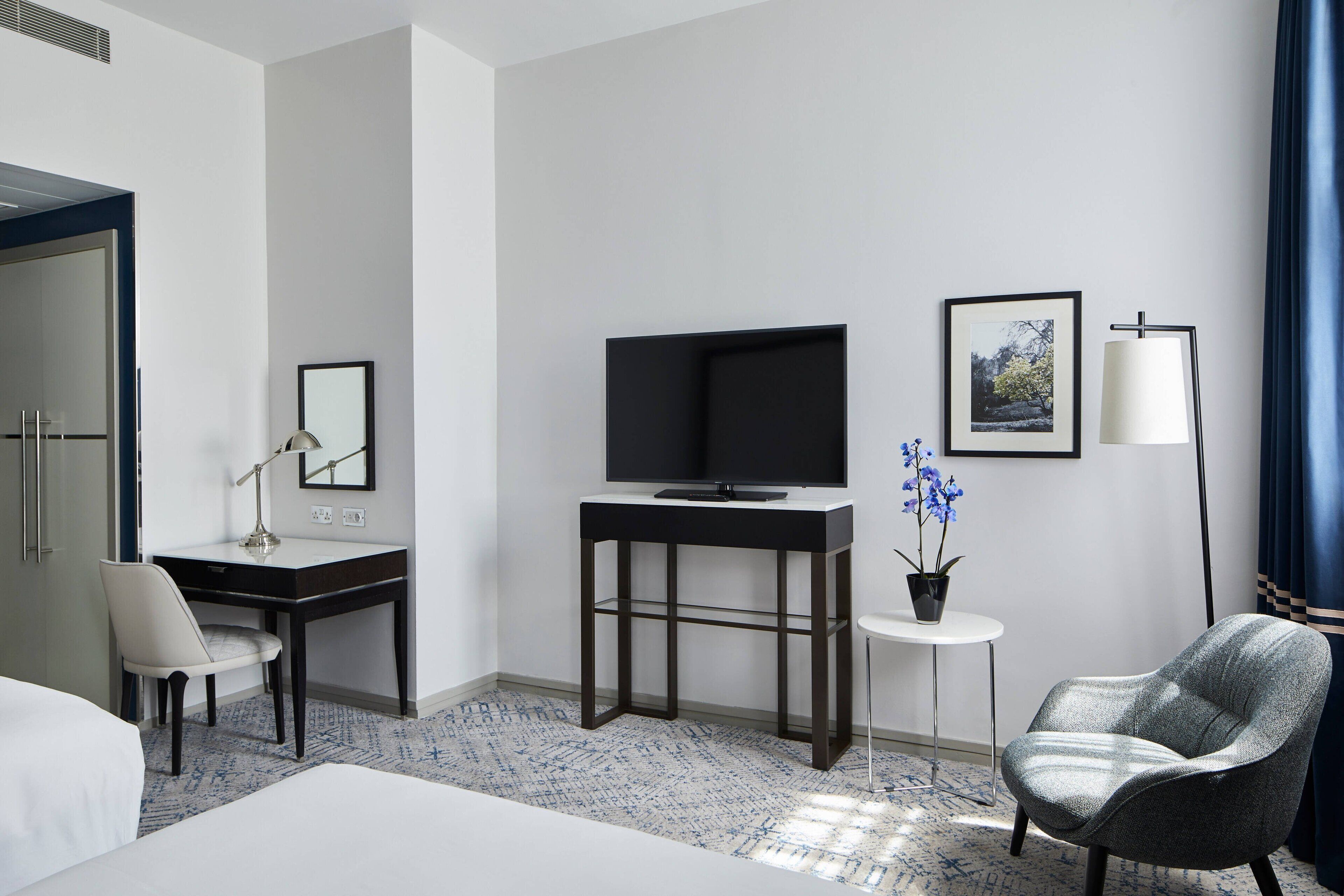 A hotel room with a bed, a desk with a chair, a television, a small table with a potted plant, and a lamp.