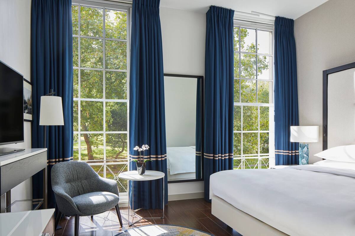 A hotel room with a bed, a desk with a TV, a chair, a small table with a vase of flowers, and large windows with blue curtains.