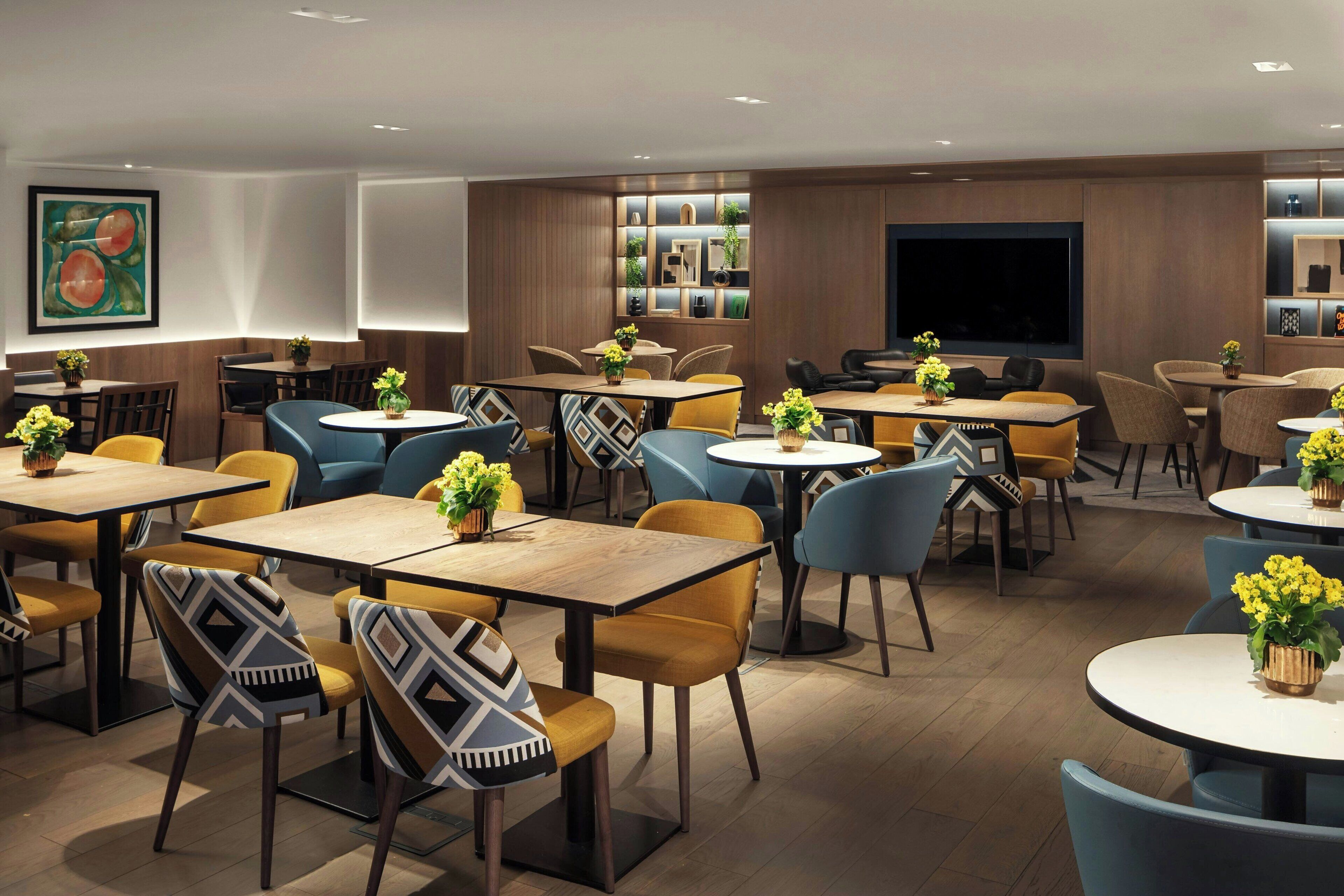 A modern dining area with multiple tables and chairs, some with yellow cushions, and a large flat-screen TV mounted on the wall.