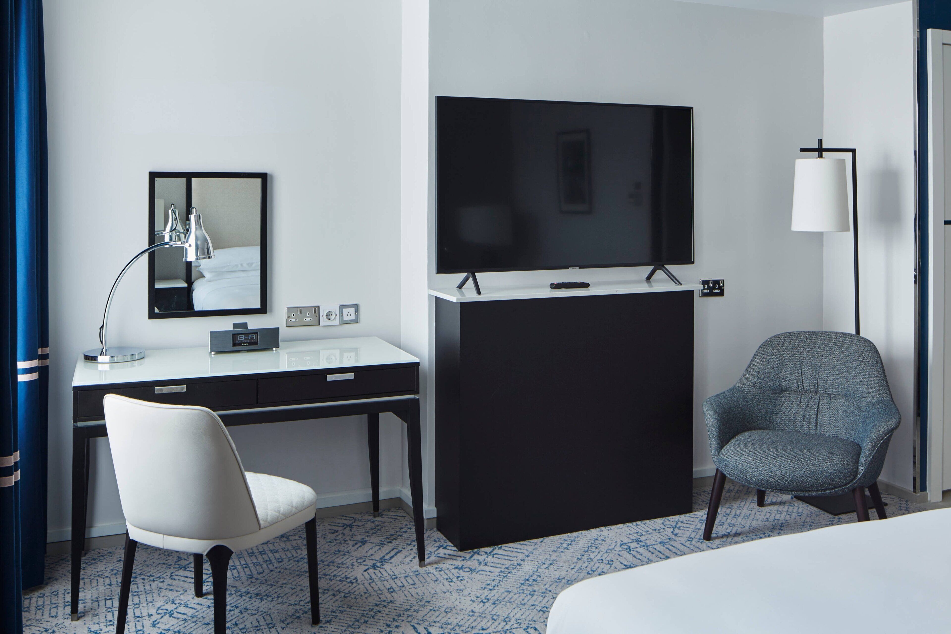 A hotel room with a bed, a desk with a chair, a television, a mirror, and a lamp.
