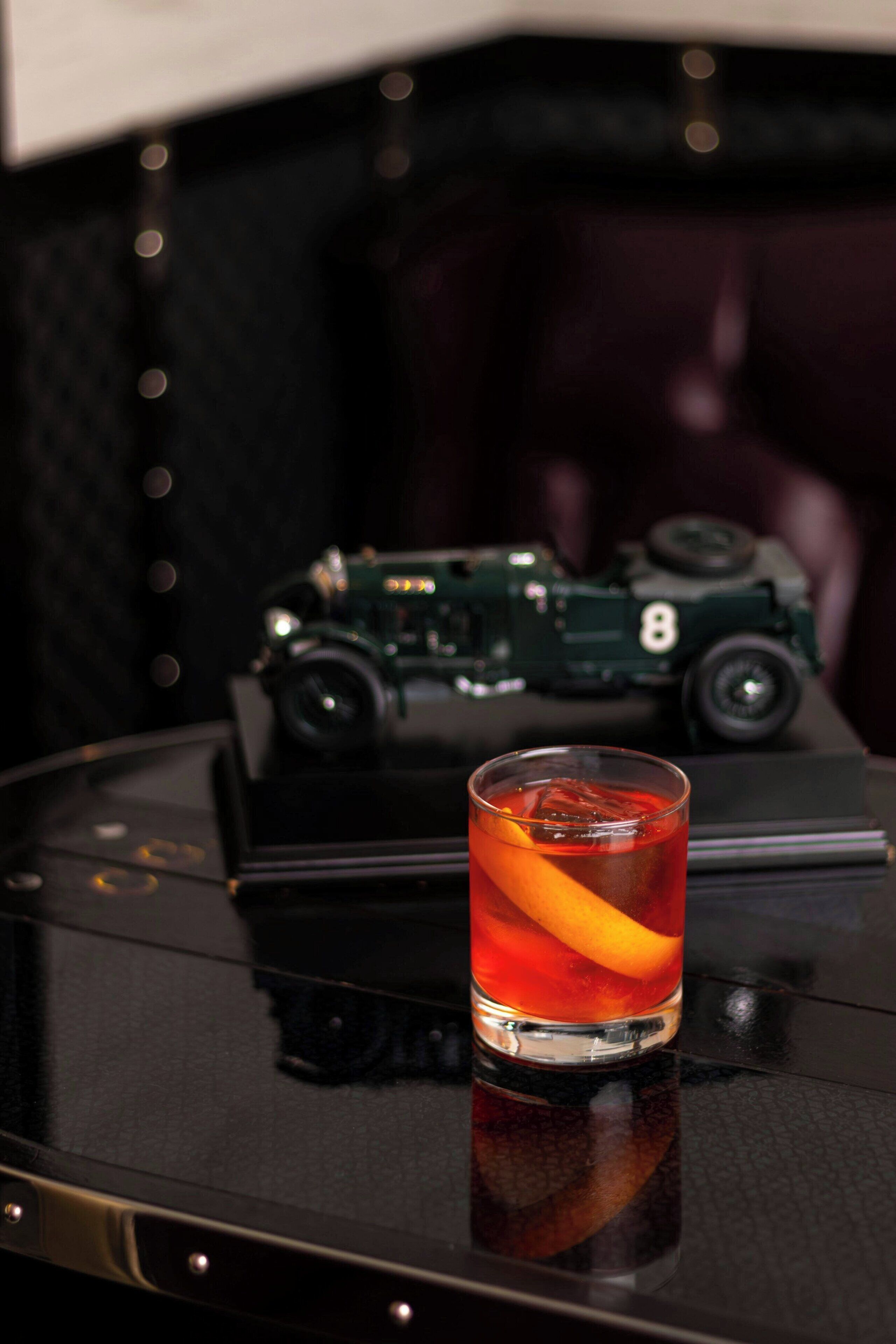 A glass of red beverage with an orange slice on a black table. A vintage green car model is in the background.