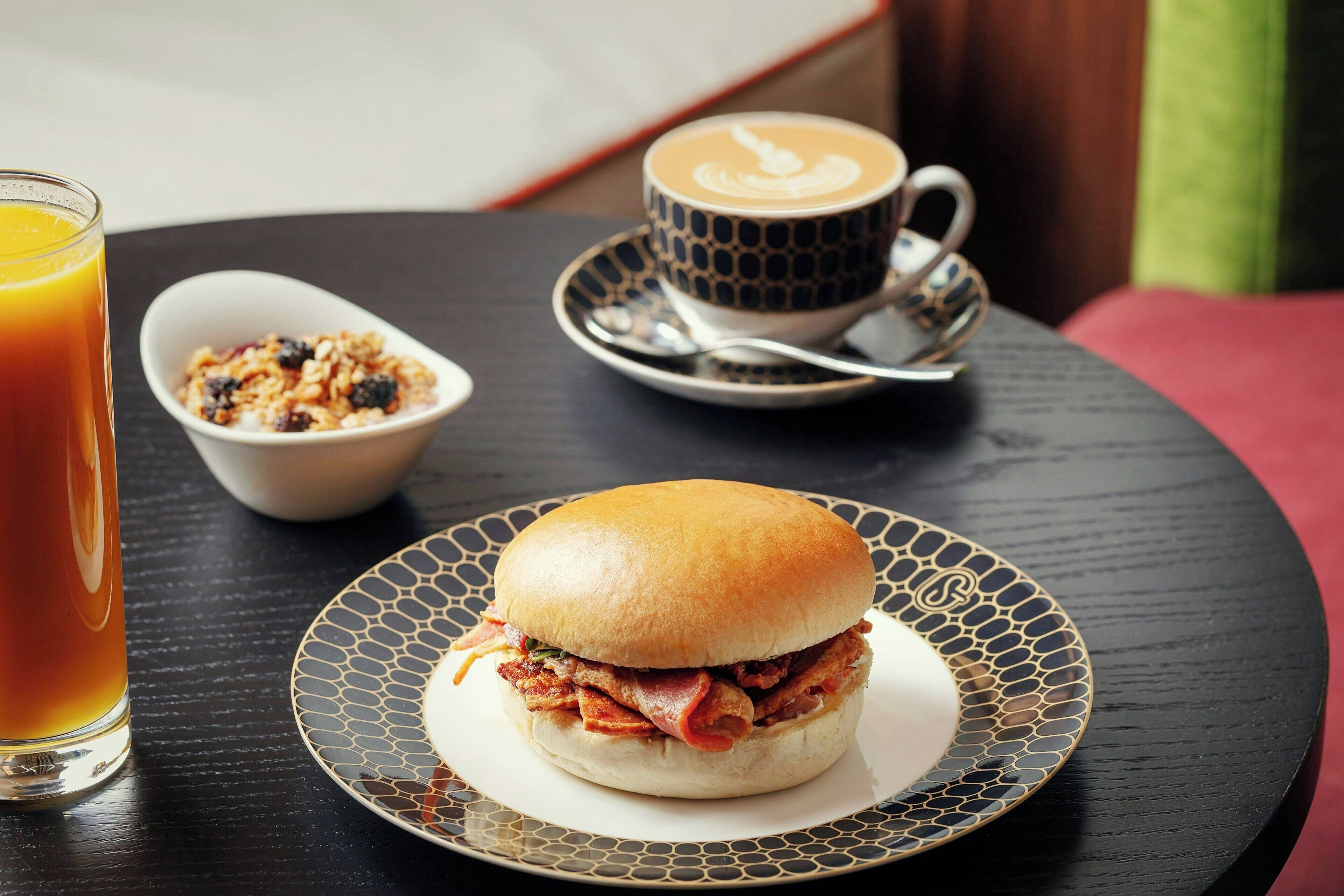 Un sándwich sobre un plato estampado, una taza de café y un bol de granola sobre una mesa.