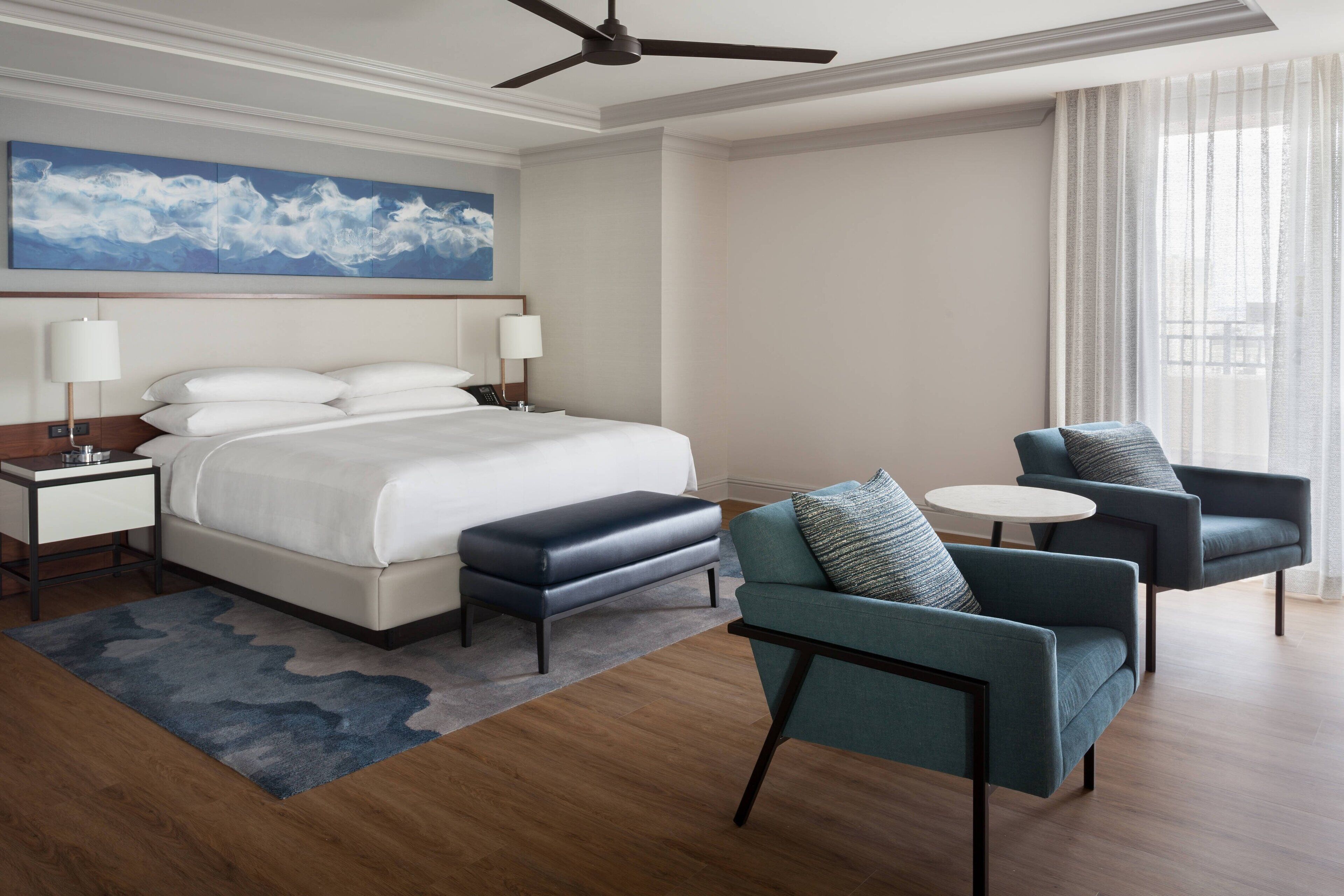 Une chambre d’hôtel comprenant un lit, deux fauteuils, une petite table, un ventilateur de plafond et une grande fenêtre avec des rideaux.