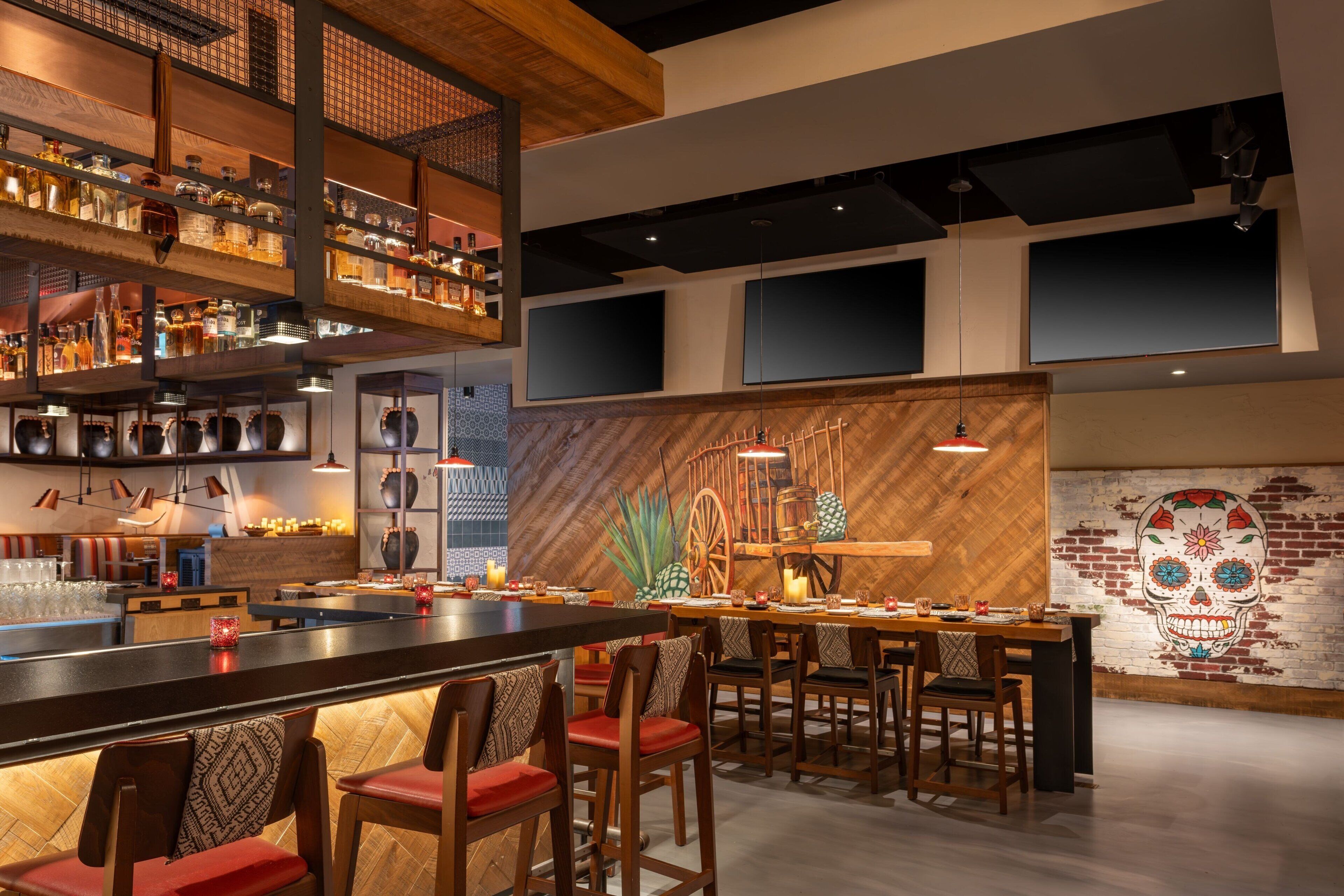 A modern bar area with multiple seating options, a variety of bottles and glasses on shelves, and a large mural on the wall.