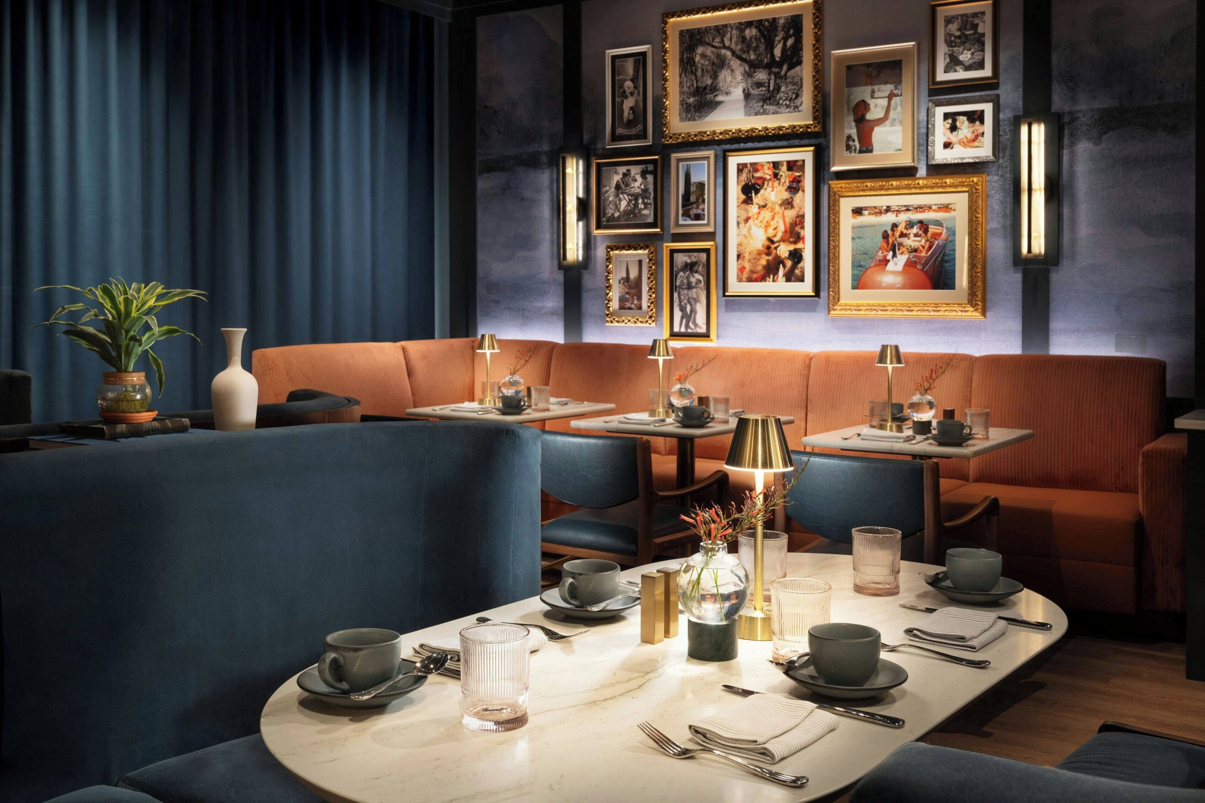 Un restaurant aux couleurs bleu et orange, avec un espace repas comprenant des tables pour deux personnes, un canapé et un mur décoré de tableaux encadrés.