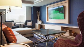 Ein modernes Wohnzimmer mit einer Couch, einem Sessel und einem Glastisch. Ein Flachbildfernseher ist an der Wand befestigt und eine Lampe ist sichtbar.
