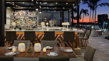 An outdoor bar area with a wooden table set for dining, wicker chairs, and a variety of bottles and glasses on shelves.