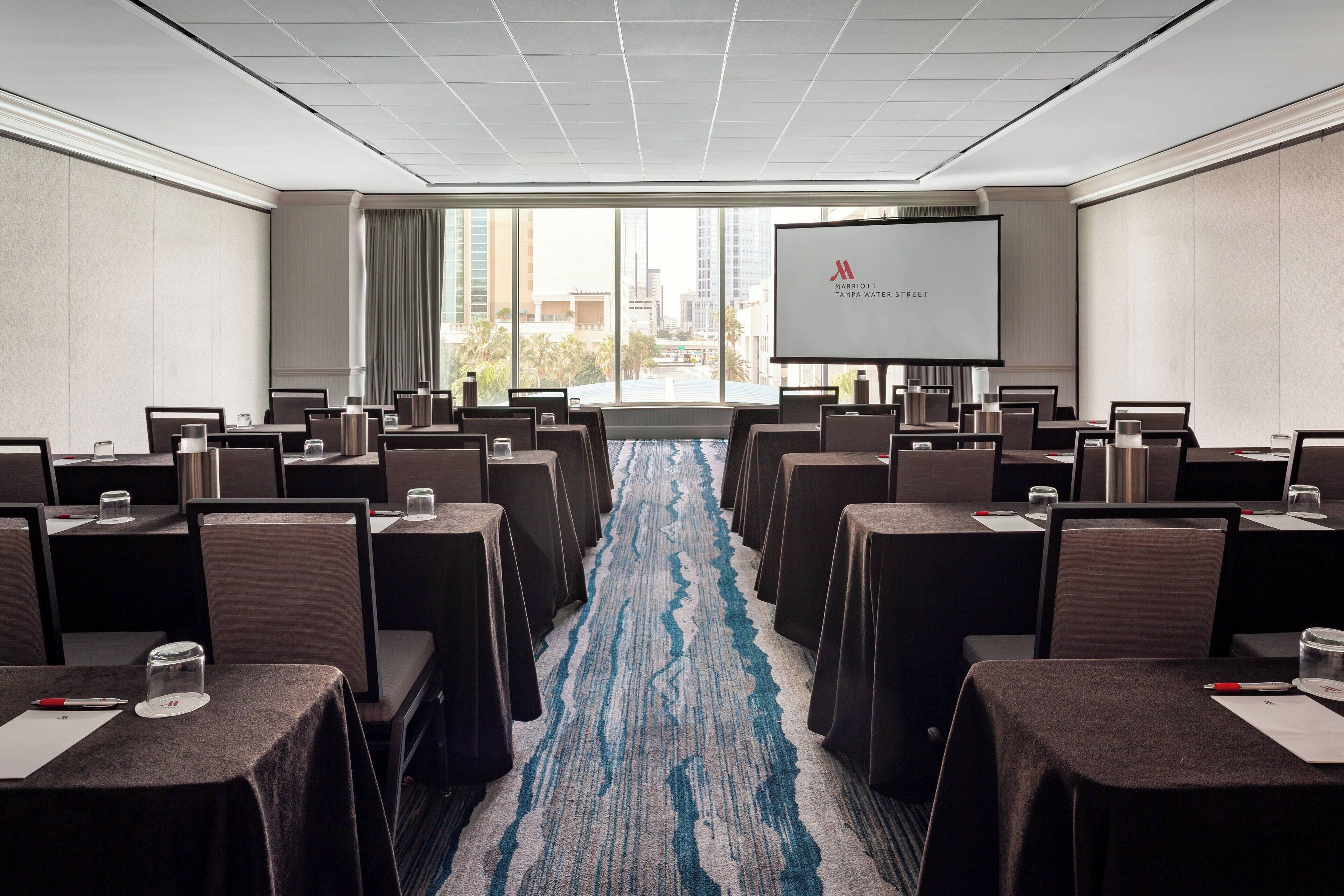 A conference room with rows of tables and chairs, a large screen at the front, and a view of the city through large windows.