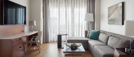 Una sala de estar moderna con televisor de pantalla plana, un escritorio con cajones, un sofá con cojín, una mesa de centro y un ventanal con cortinas traslúcidas.