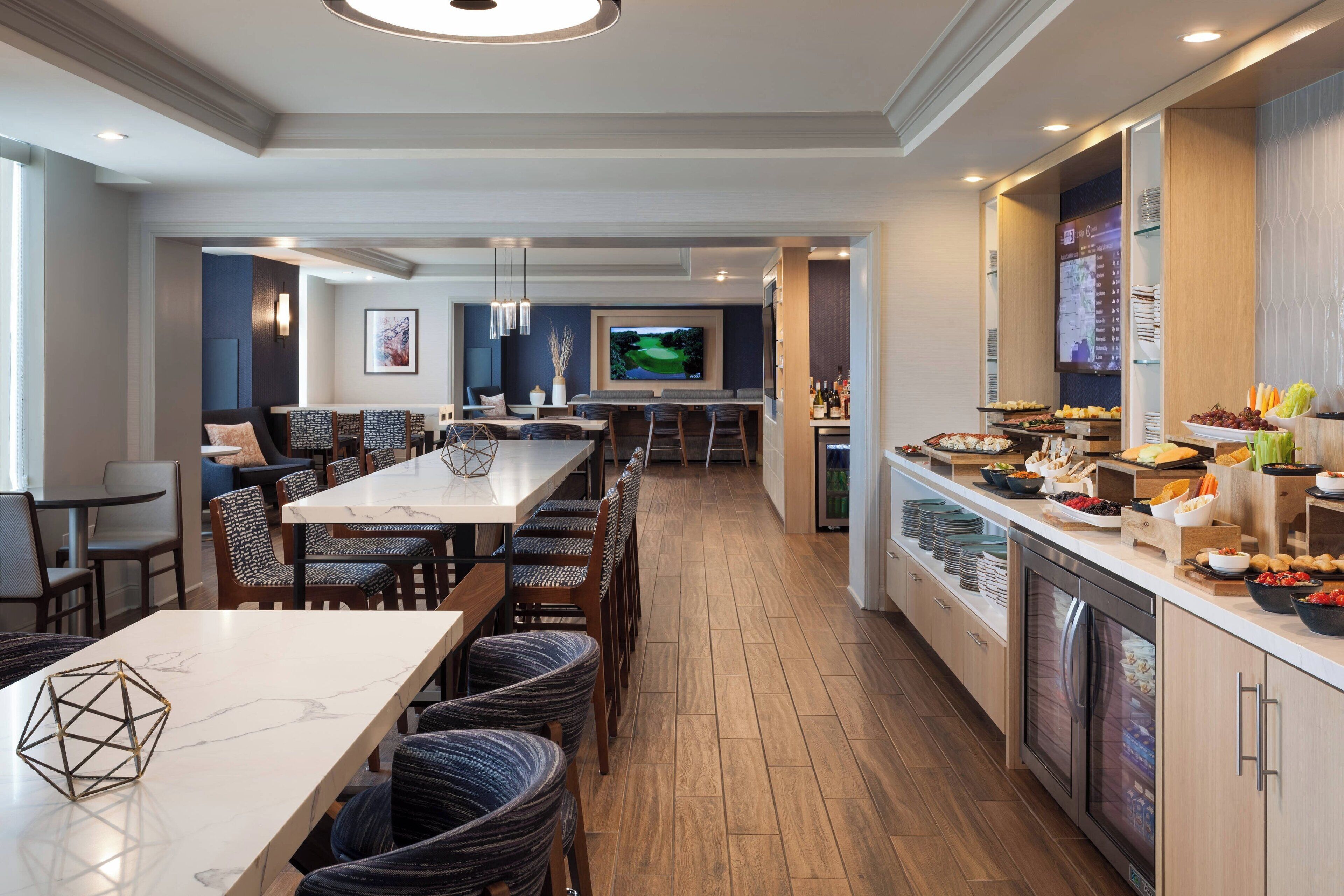 A modern hotel dining area with a long communal table, comfortable seating, a television, and a buffet with various food items.