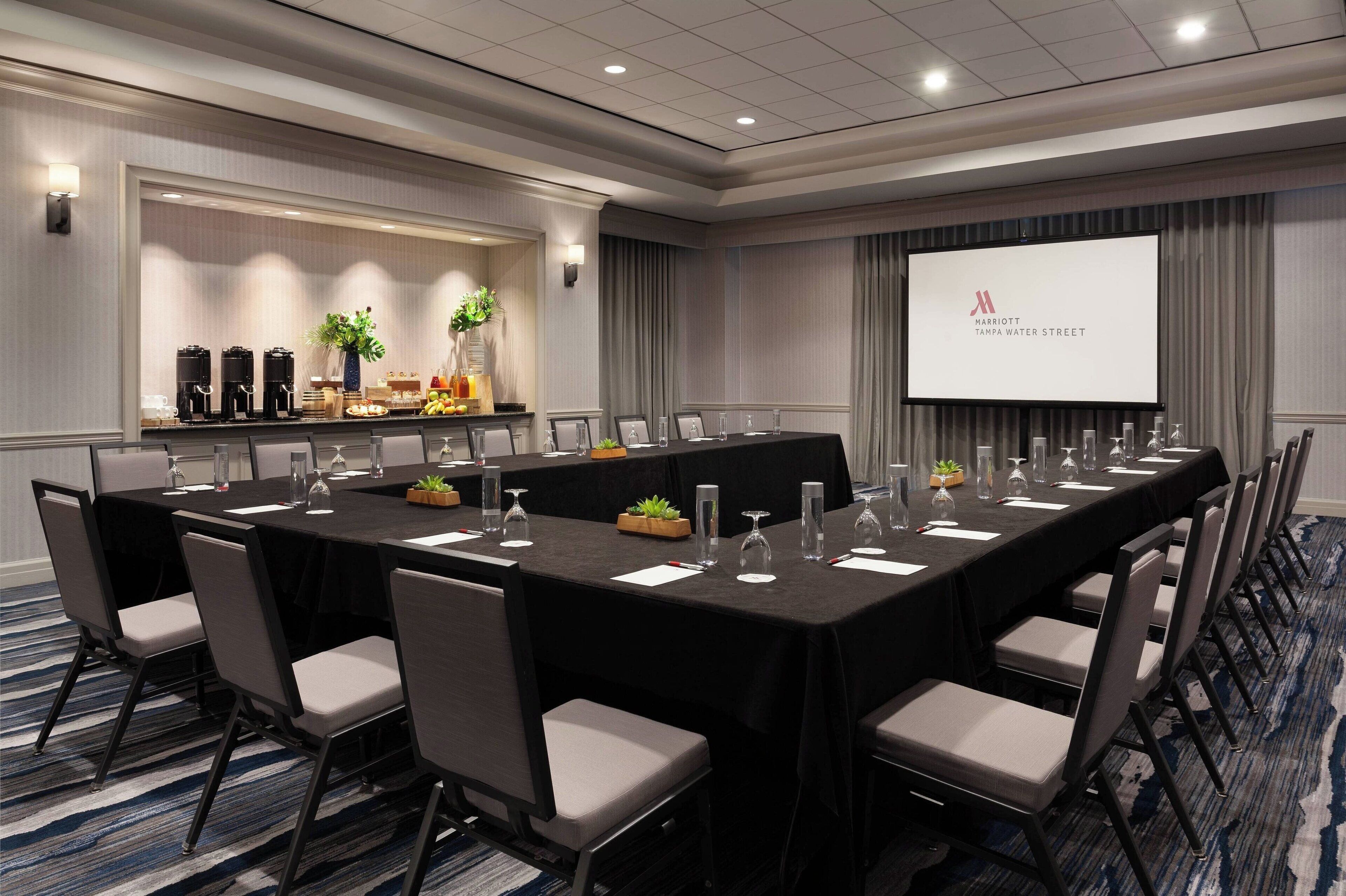 Un salón de conferencias con una larga mesa, sillas, una pantalla de proyector y una zona de buffet con cafeteras y fruta.