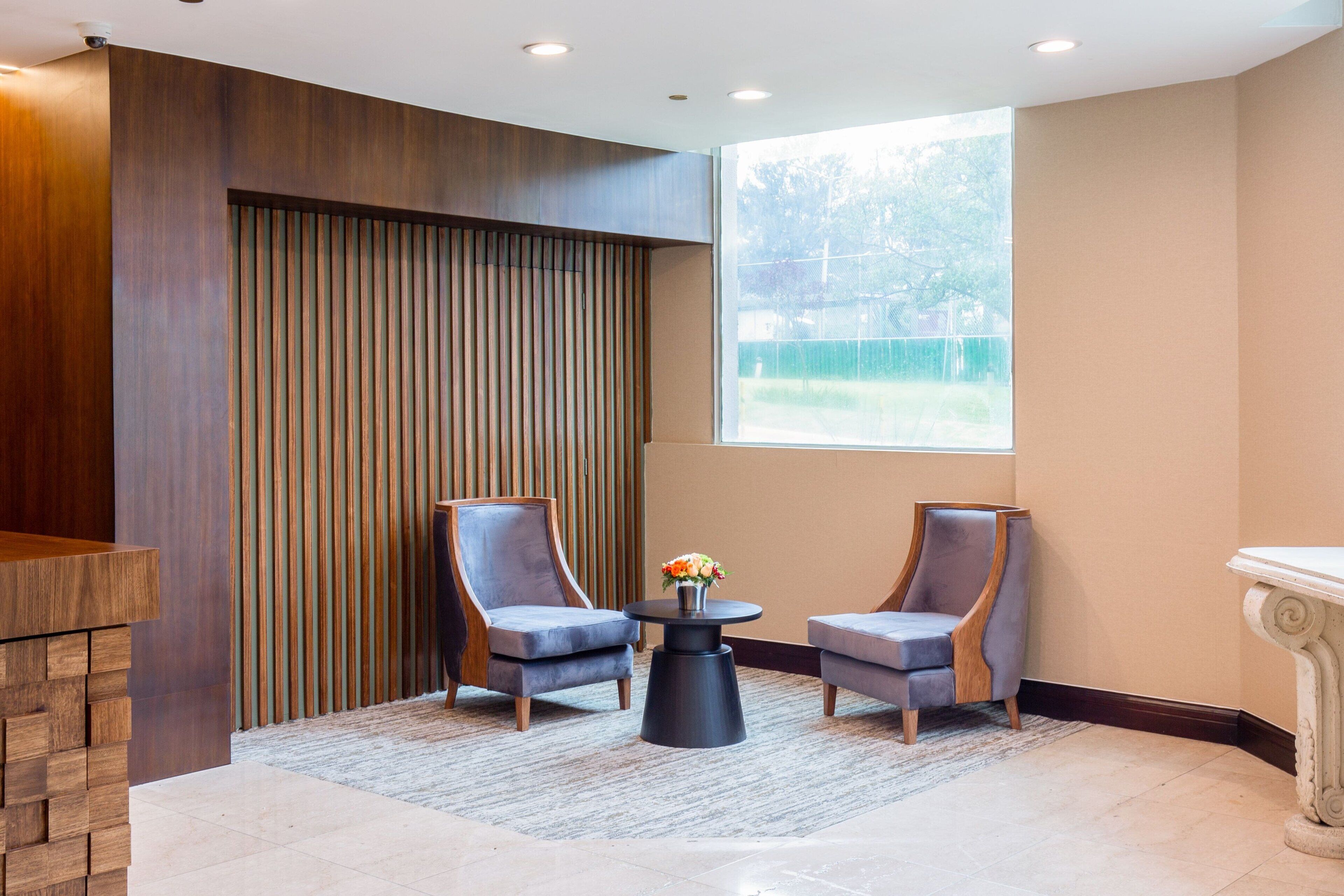 A modern lobby with two grey armchairs, a small round table with a vase of flowers, and a wooden reception desk.