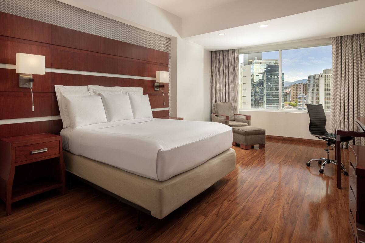 A hotel room with a large bed, wooden headboard, two bedside lamps, a desk with a chair, and a window with a city view.
