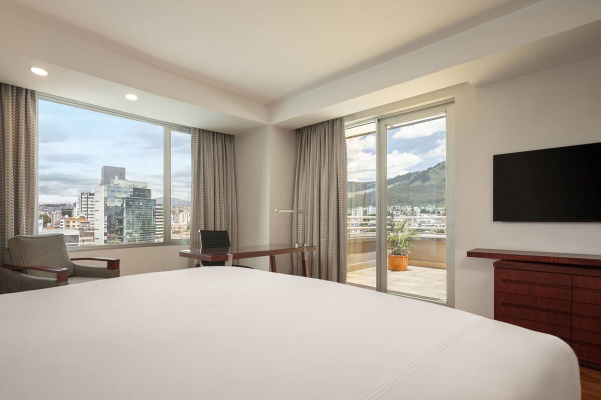 A hotel room with a large bed, a desk, a chair, a TV, and a balcony with a city view.