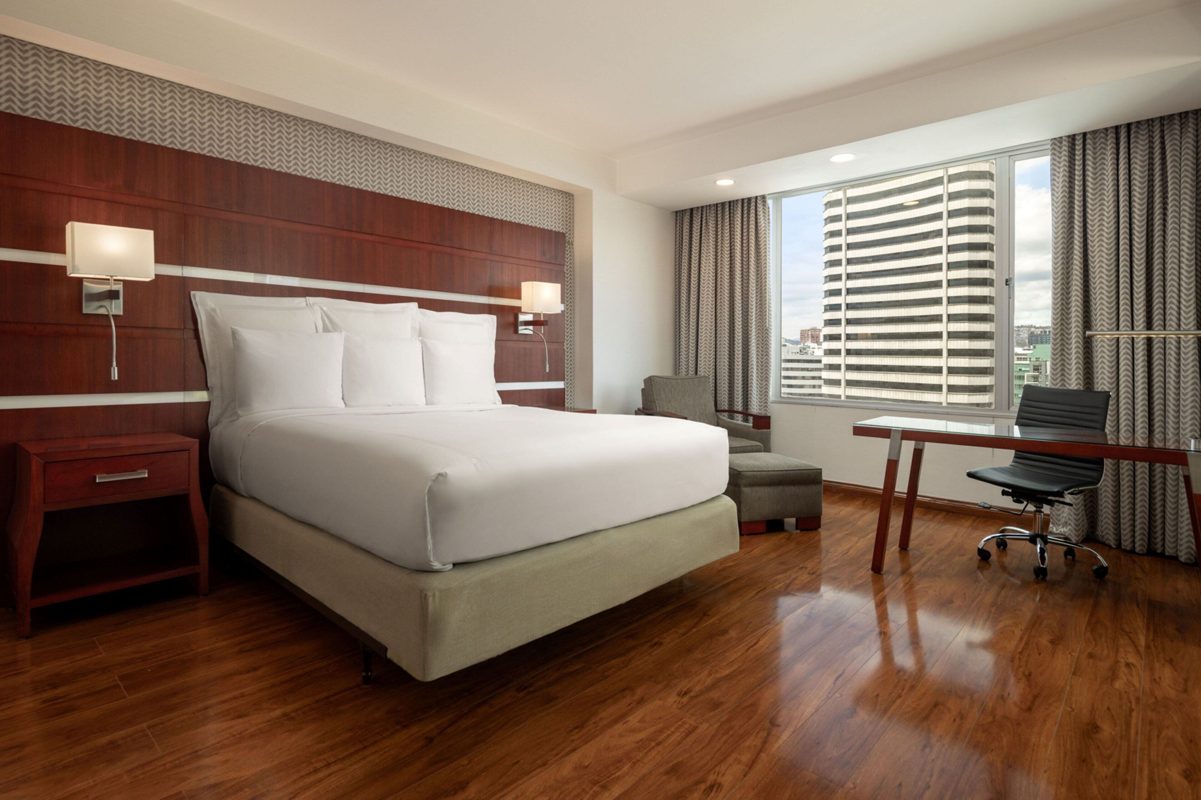 Quarto de hotel com uma cama grande, uma escrivaninha, uma cadeira e vista para a cidade pela janela.