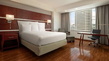 Quarto de hotel com uma cama grande, uma escrivaninha, uma cadeira e vista para a cidade pela janela.