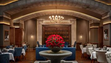 Une salle à manger élégante, ornée d’un lustre, d’un grand arrangement floral et d’une fontaine en pierre. Les tables sont dressées avec des nappes blanches et des chaises bleues.