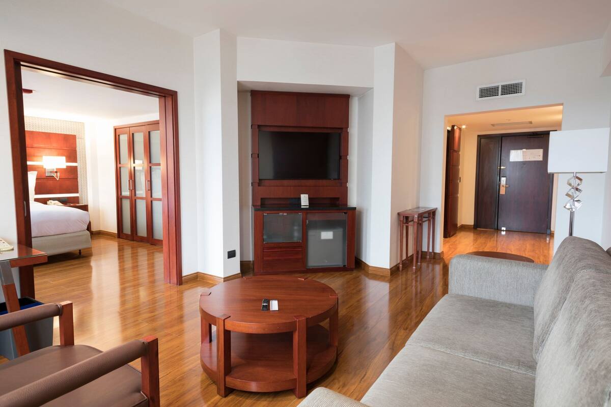 A hotel room with a wooden floor, a sofa, a round table, a TV, a minibar, and a door leading to another room.
