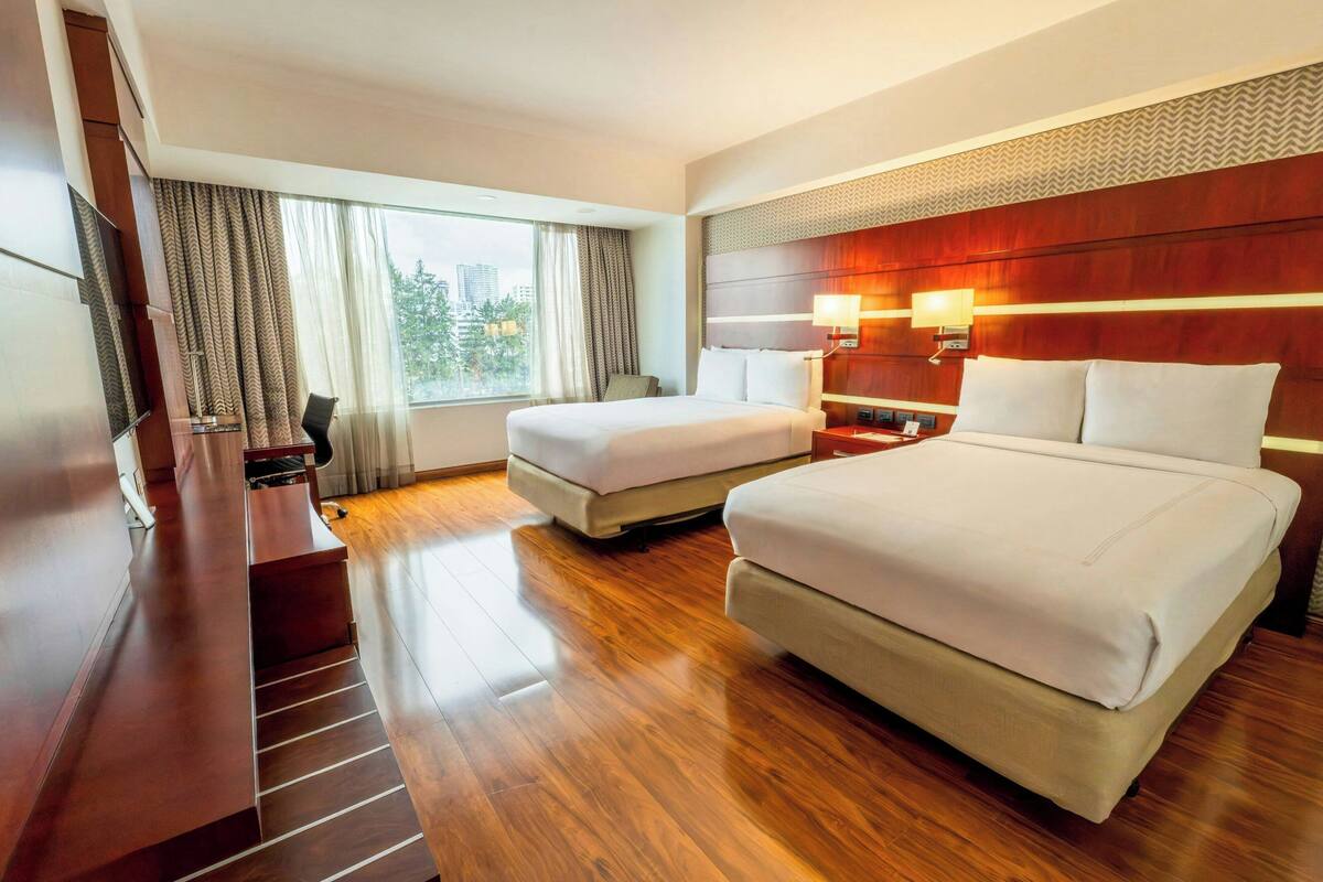 A hotel room with a large bed, a smaller bed, a desk, a chair, a TV, and a window with curtains.