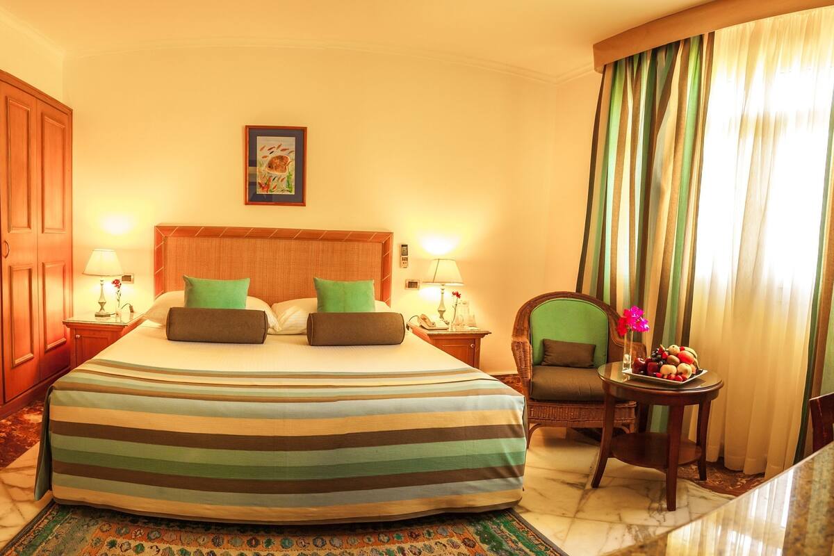 A hotel room with a large bed, two bedside tables with lamps, a chair, a small table with fruit, and a large wardrobe.