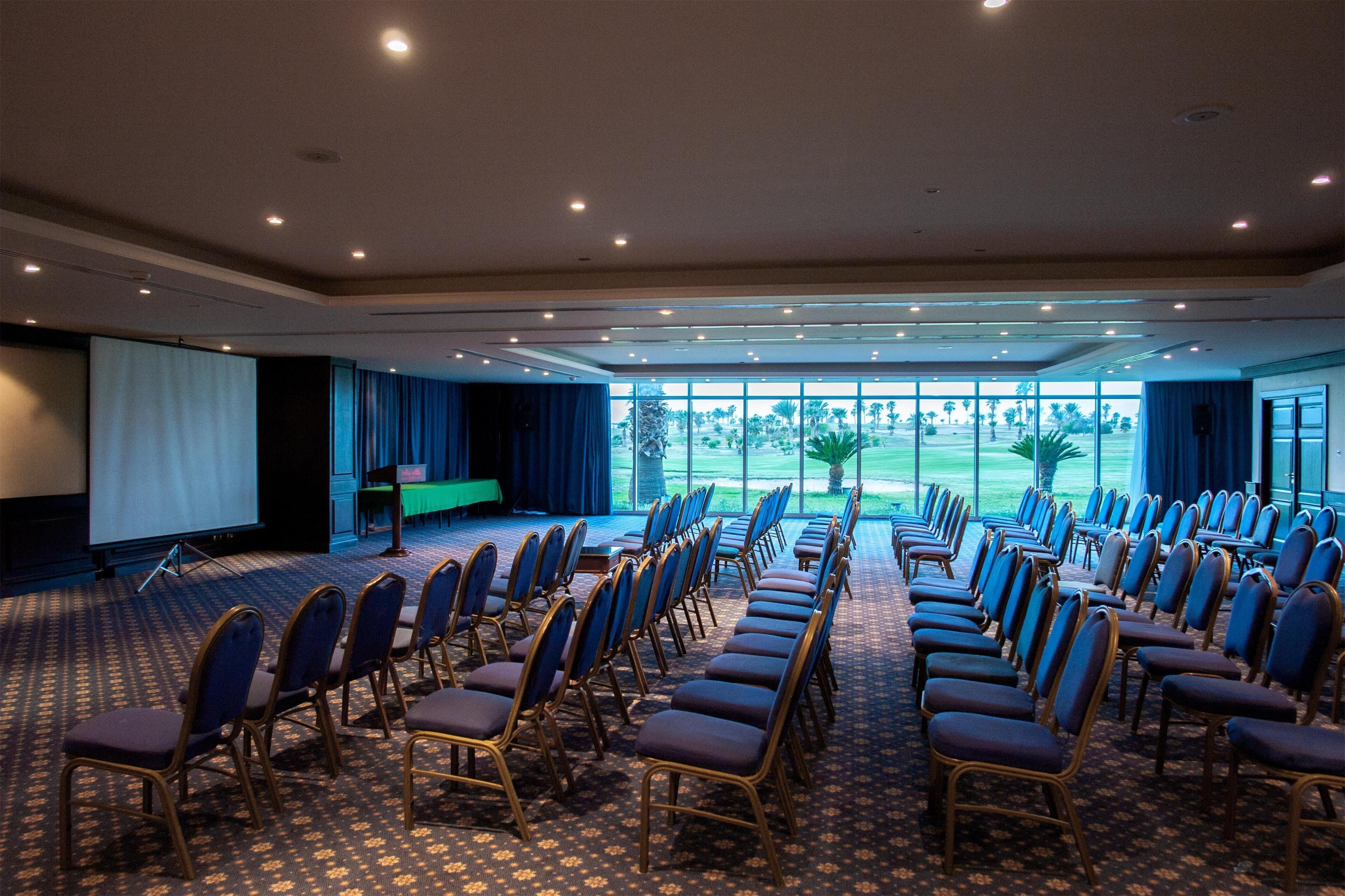 Une salle de conférence avec des rangées de chaises, un pupitre et un grand écran.