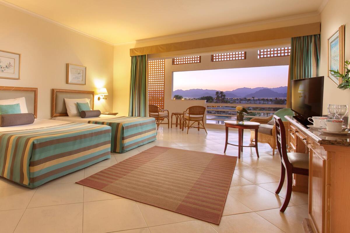A hotel room with two beds, a TV, a desk with a chair, and a view of the cityscape.