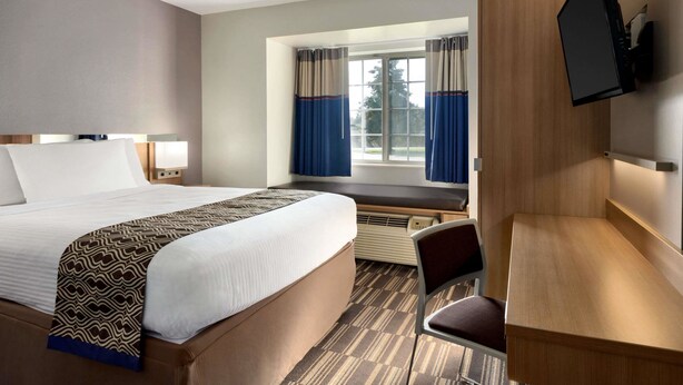 A hotel room with a bed, a desk, a chair, a TV mounted on the wall, and a window with curtains. - Microtel Inn & Suites by Wyndham Baton Rouge (Baton Rouge)