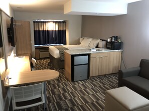 A compact hotel room with a kitchenette, a bed with a window, a small desk, and a seating area.