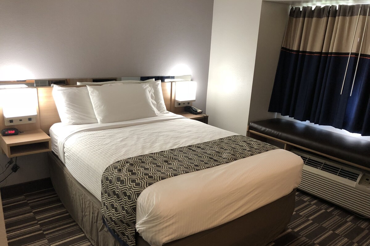 A hotel room with a bed, bedside lamps, a nightstand, a window with curtains, and an air conditioning unit.