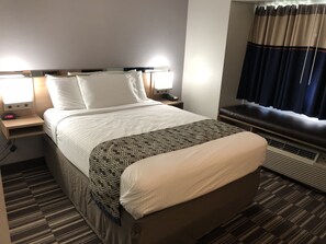 A hotel room with a bed, bedside lamps, a nightstand, a window with curtains, and an air conditioning unit.