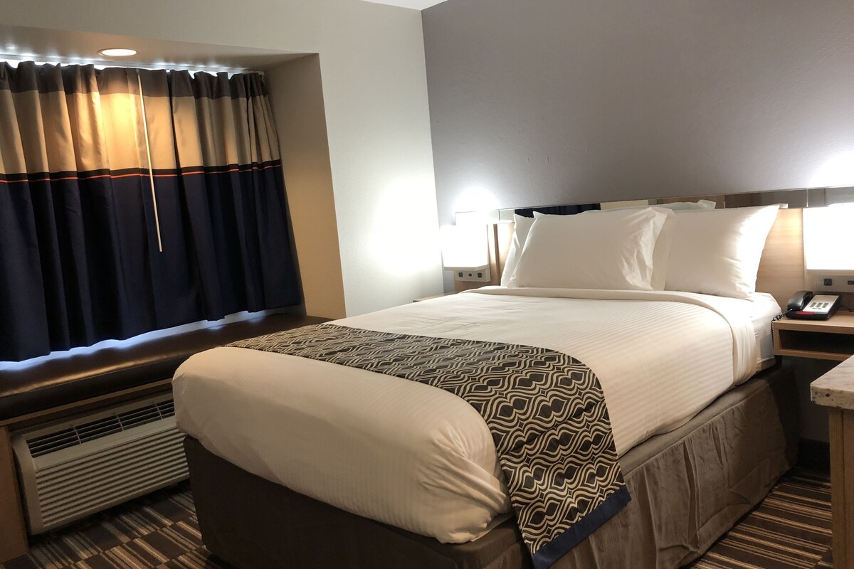A hotel room with a large bed, a bedside table with a phone, a heater, and a window with curtains.