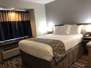 A hotel room with a large bed, a bedside table with a phone, a heater, and a window with curtains.