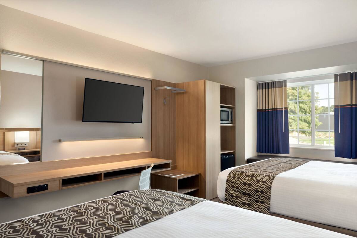 Habitación de hotel con cama, televisor montado en la pared, escritorio con silla y ventana con cortinas.