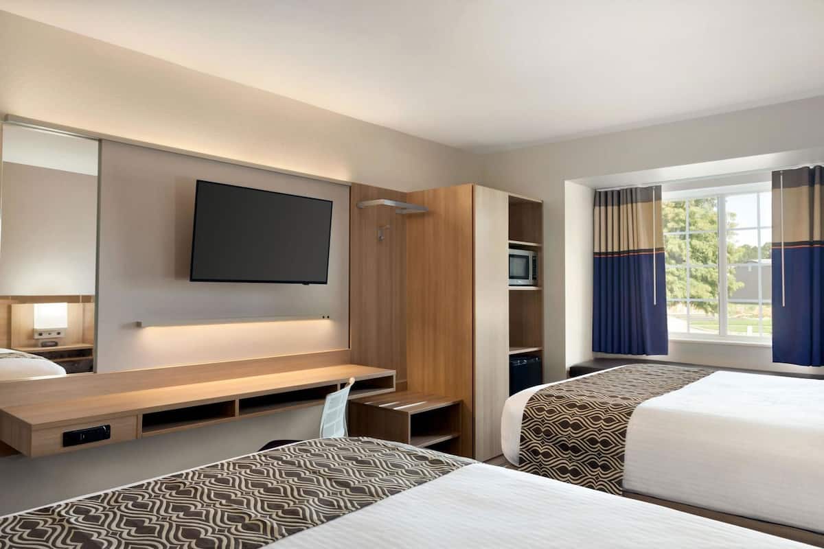 Una habitación de hotel con televisión instalada en la pared, un escritorio de madera, microondas y ventana con cortinas.