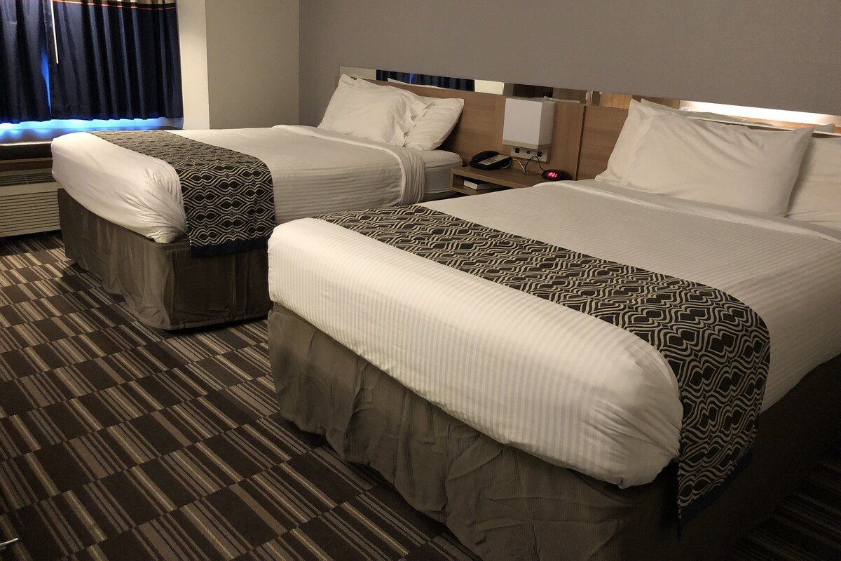 A hotel room with two beds, a bedside table with a phone and alarm clock, and a window with curtains.