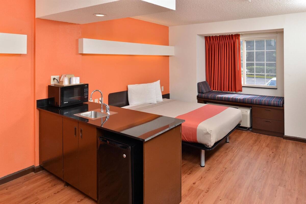 Een compacte hotelkamer met kitchenette, een bed, een raam met gordijnen en een zithoek bij het raam.