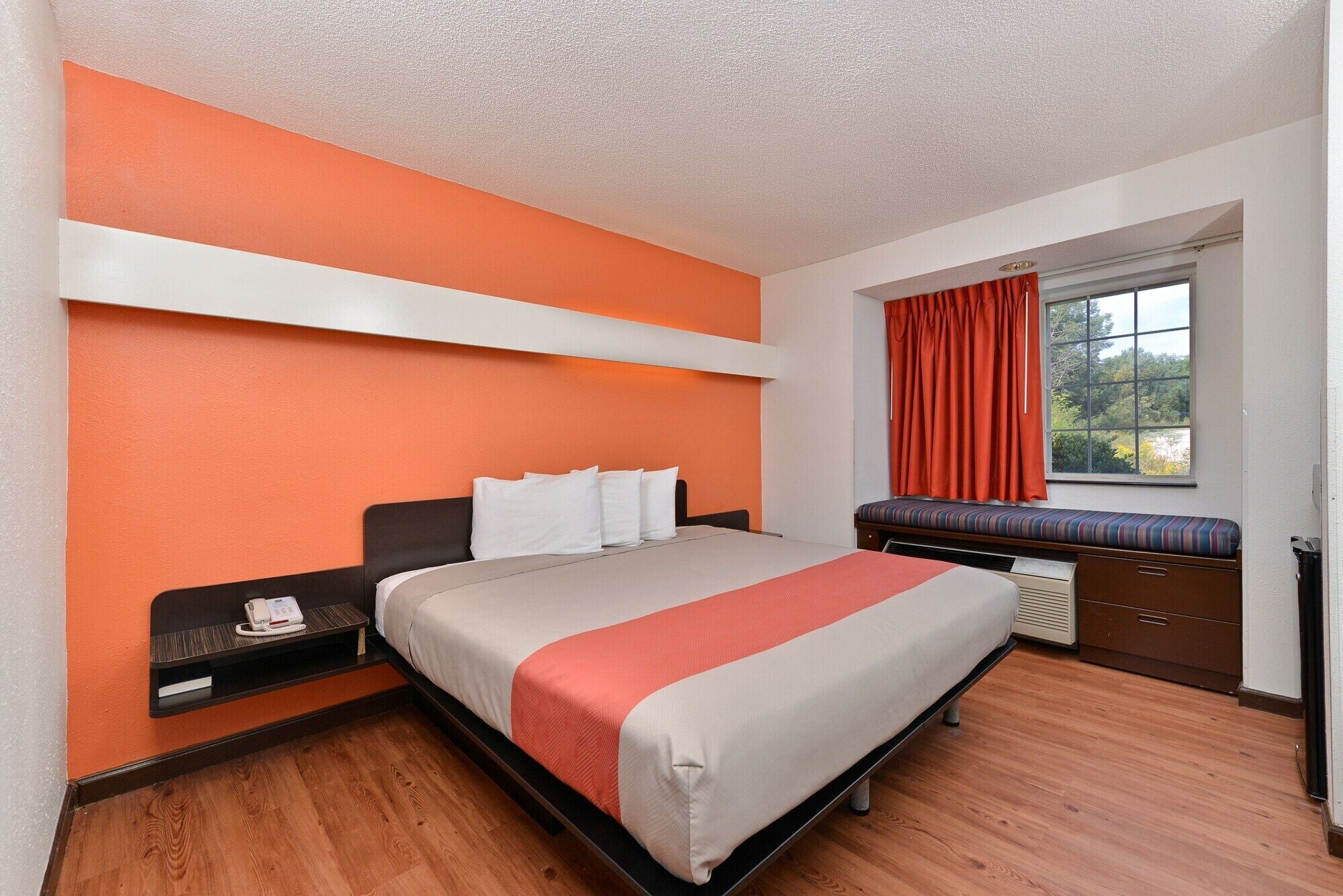 A hotel room with a bed, a bedside table with a phone, a window with curtains, and a view of trees.