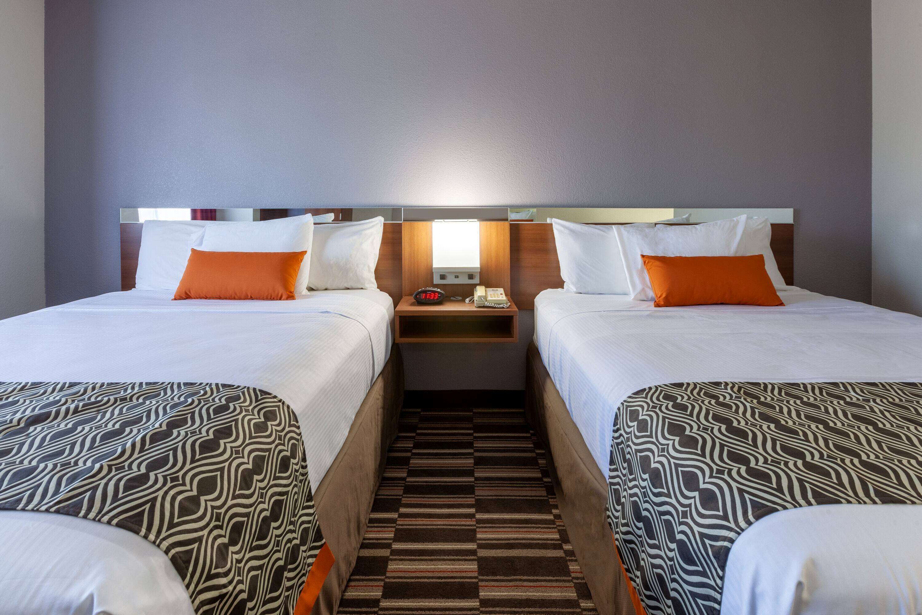 Une chambre d’hôtel avec deux lits, chacun agrémenté de draps blancs et d’oreillers orange, une table de chevet avec une lampe et un tapis à rayures.
