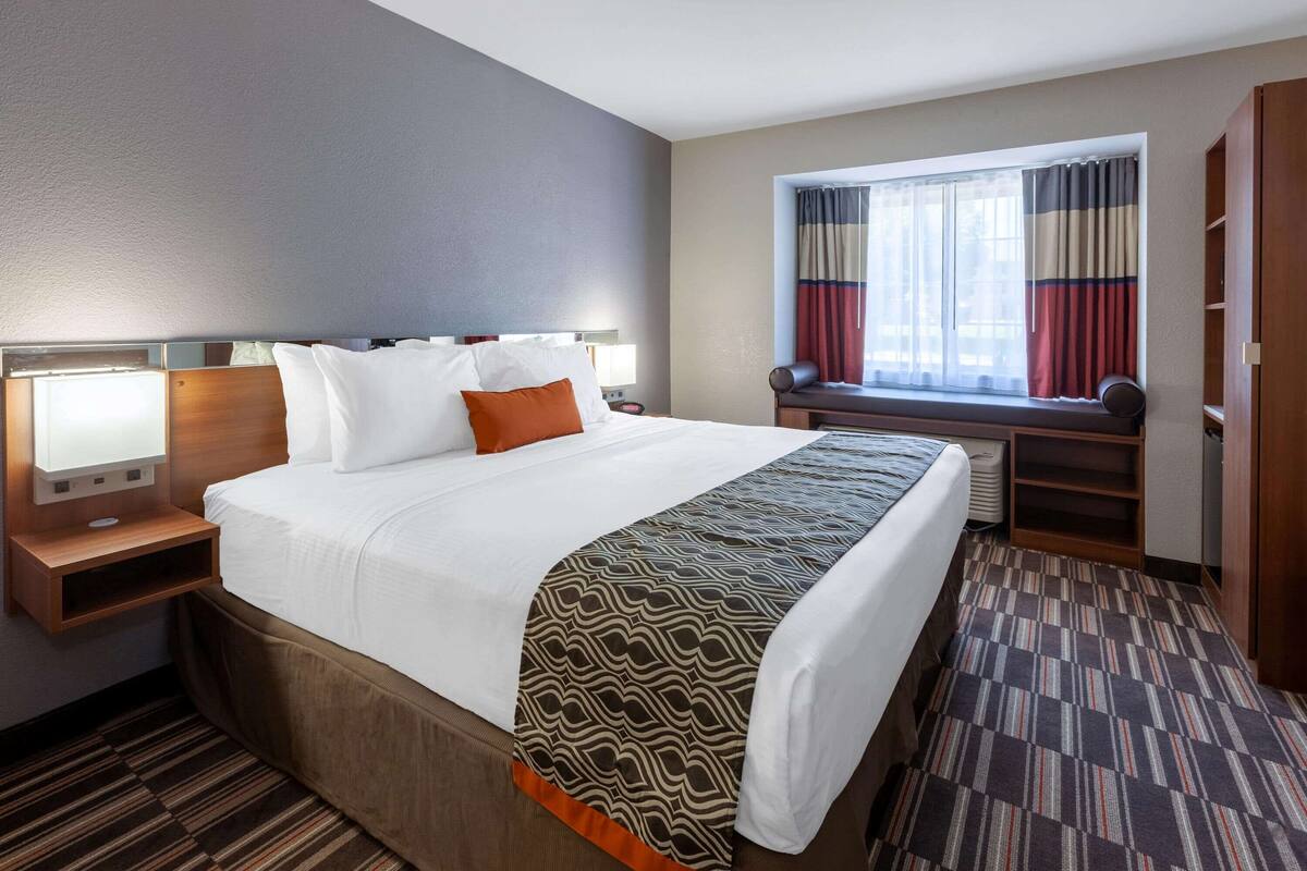 Una habitación de hotel con una cama grande, una mesita de noche con lámpara, una ventana con cortinas y un armario empotrado.