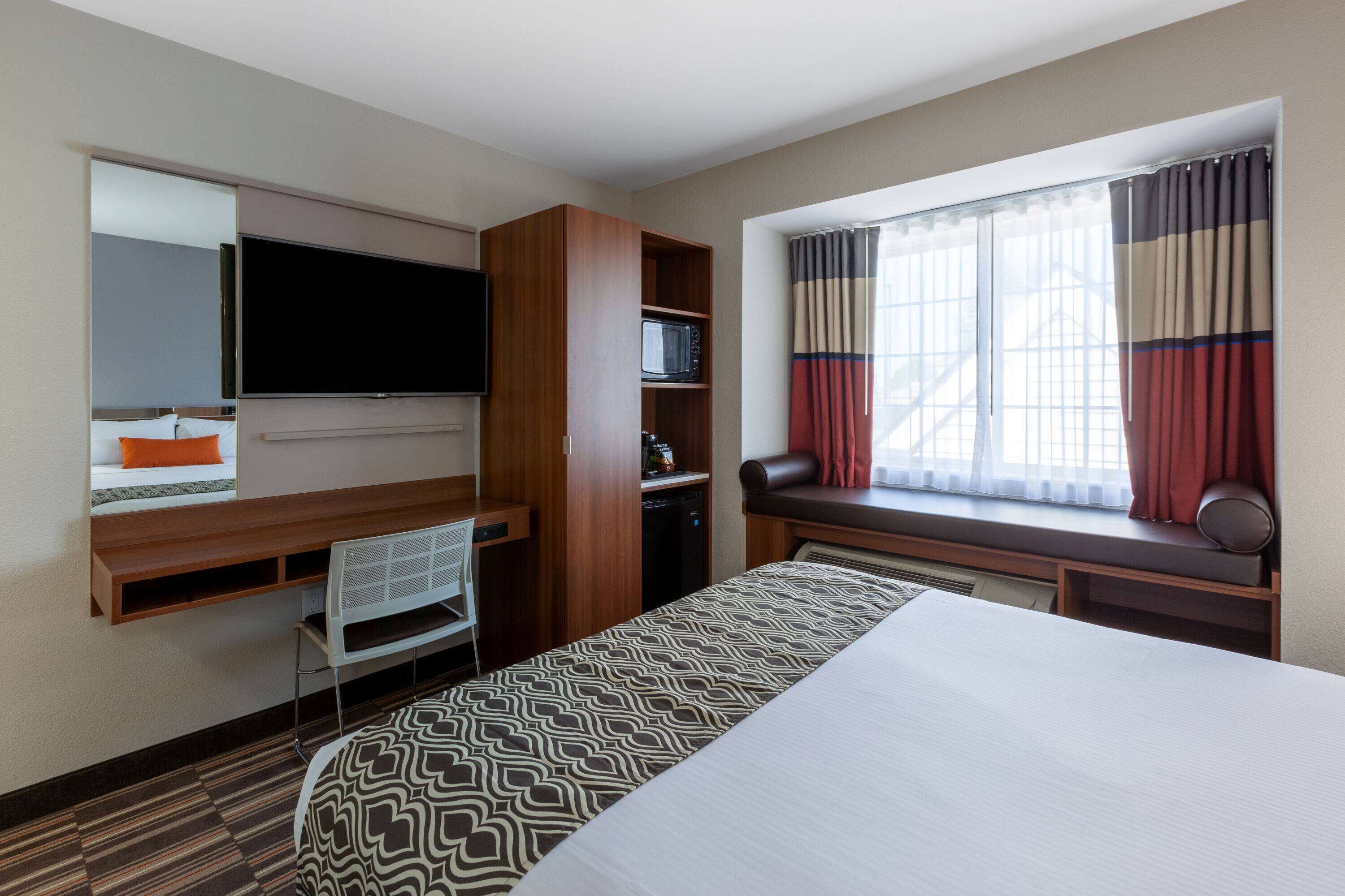 A hotel room with a bed, a TV, a desk with a chair, a microwave, and a window with curtains.