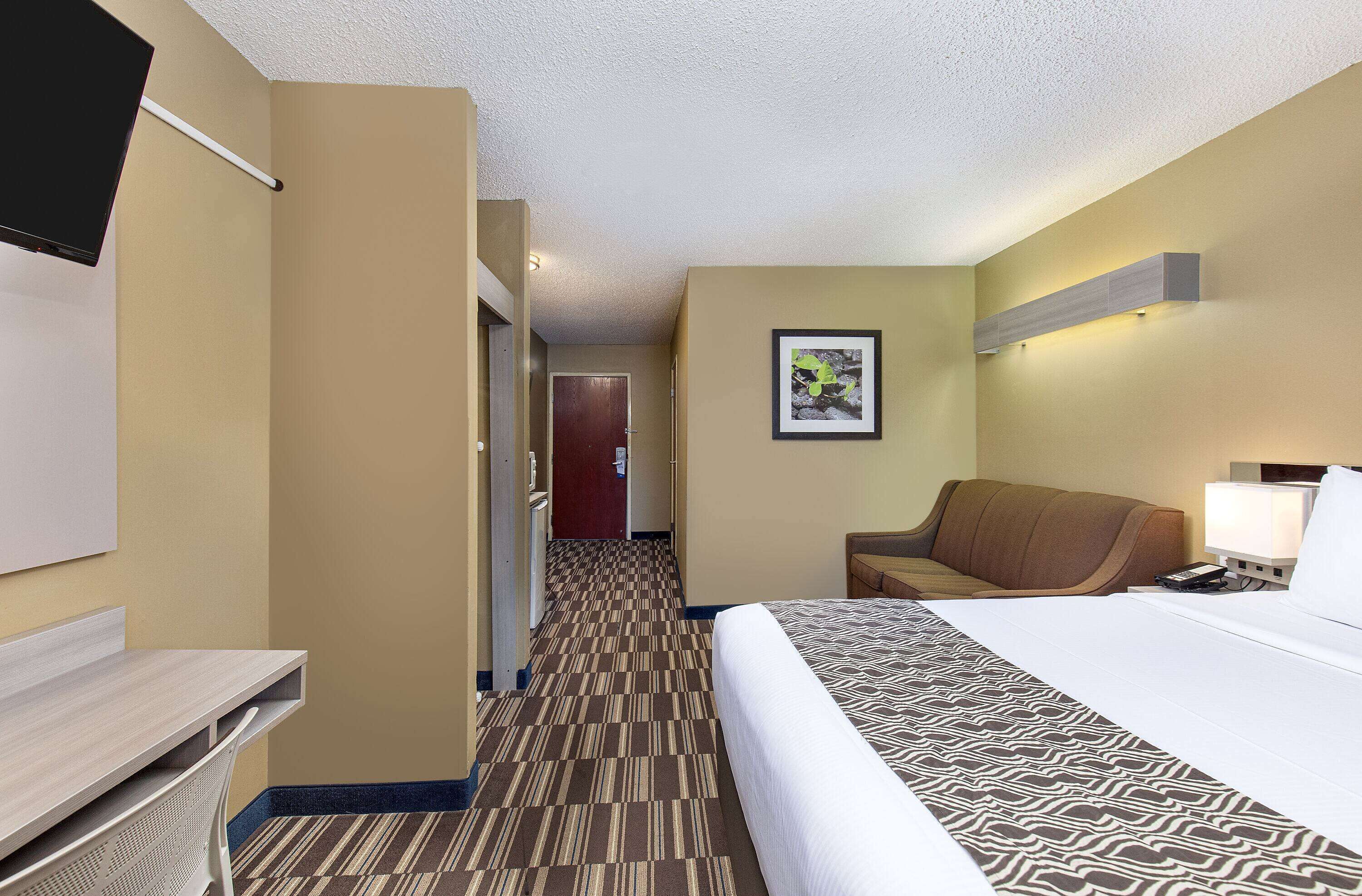 A hotel room with a bed, a desk, a chair, a sofa, a framed picture on the wall, and a hallway leading to another room.