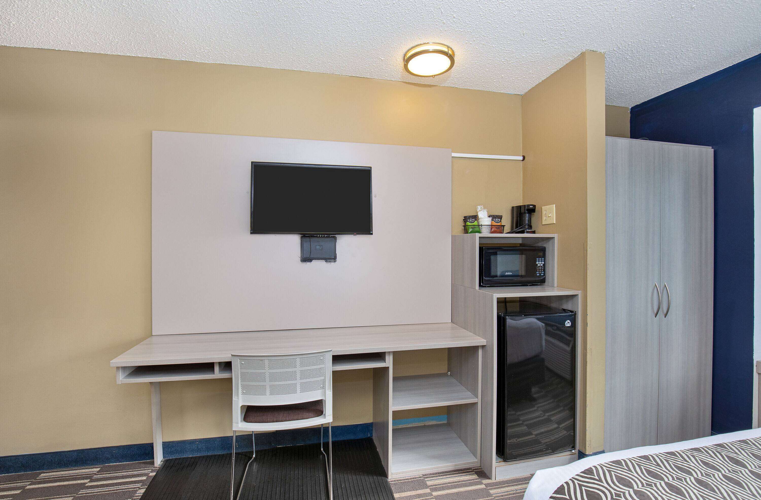 A compact hotel room with a kitchenette, a flat-screen TV mounted on the wall, a bed with a patterned bedspread, and a wardrobe.