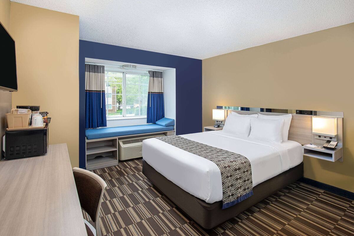 A hotel room with a large bed, a desk with a computer, a window with curtains, and a small seating area.