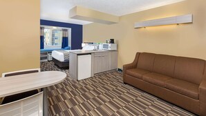 Una habitación de hotel compacta con kitchenette, un sofá marrón, una mesita y una ventana con cortinas.