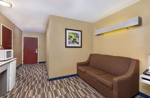 Habitación de hotel con un sofá marrón, microondas, refrigerador, un cuadro enmarcado en la pared y un pasillo con alfombra estampada.