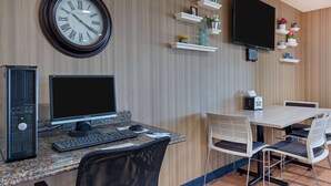 Habitación de hotel con computadora de escritorio, un reloj de pared grande, televisor de pantalla plana y un área de comedor con mesa y sillas.