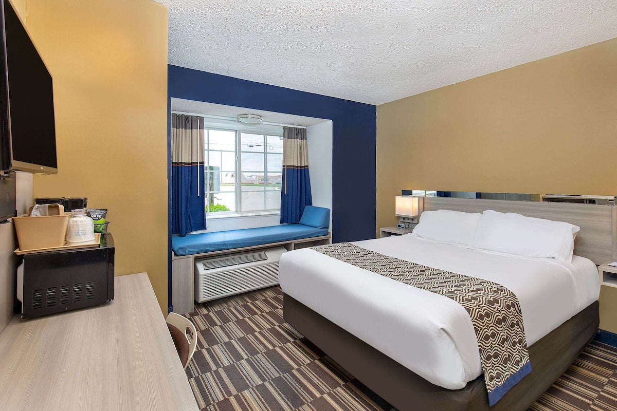 A hotel room with a large bed, a desk with a computer, a window with curtains, and a view of the outside.