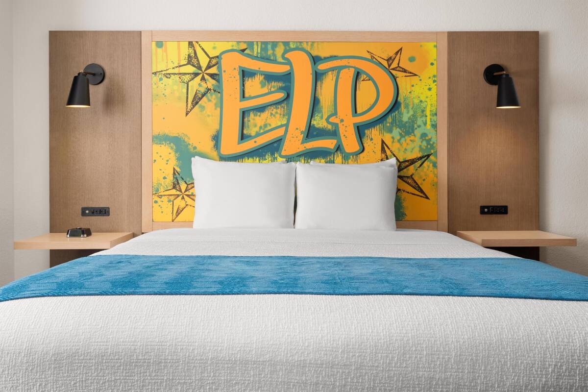 Una camera d'albergo con un letto, due comodini, una lampada a parete e un'opera d'arte colorata sopra il letto.